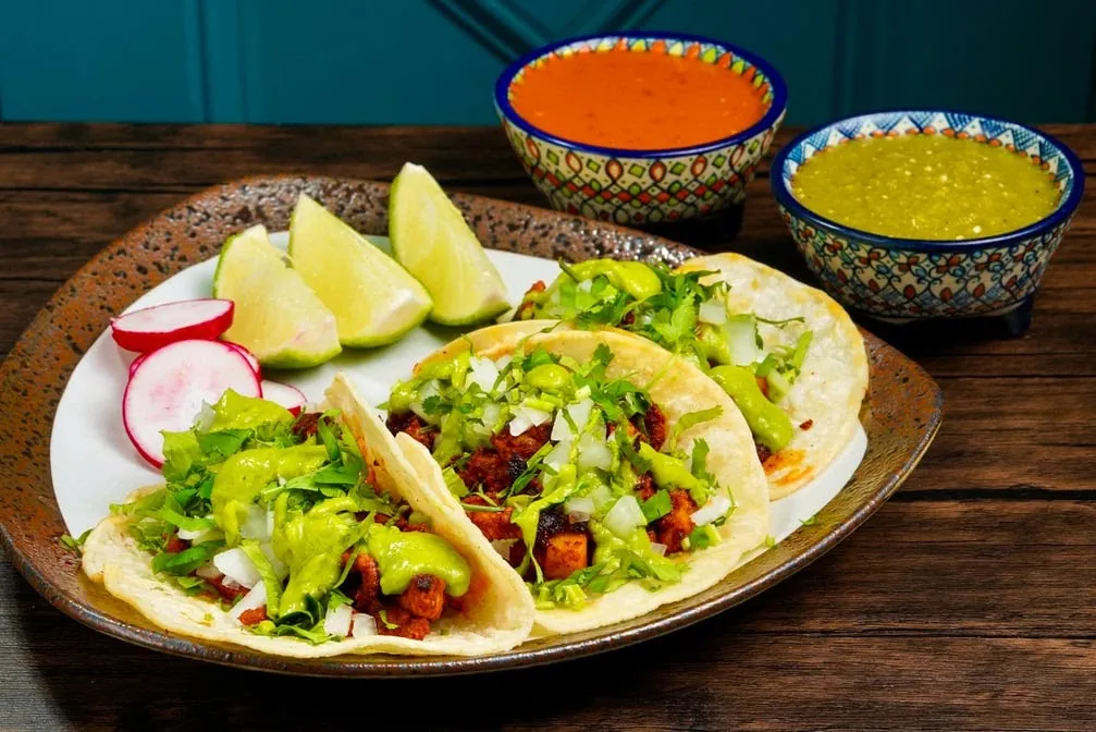 Three al pastor tacos with avocado salsa, onion and cilantro, with lime, radishes and red and green salsas