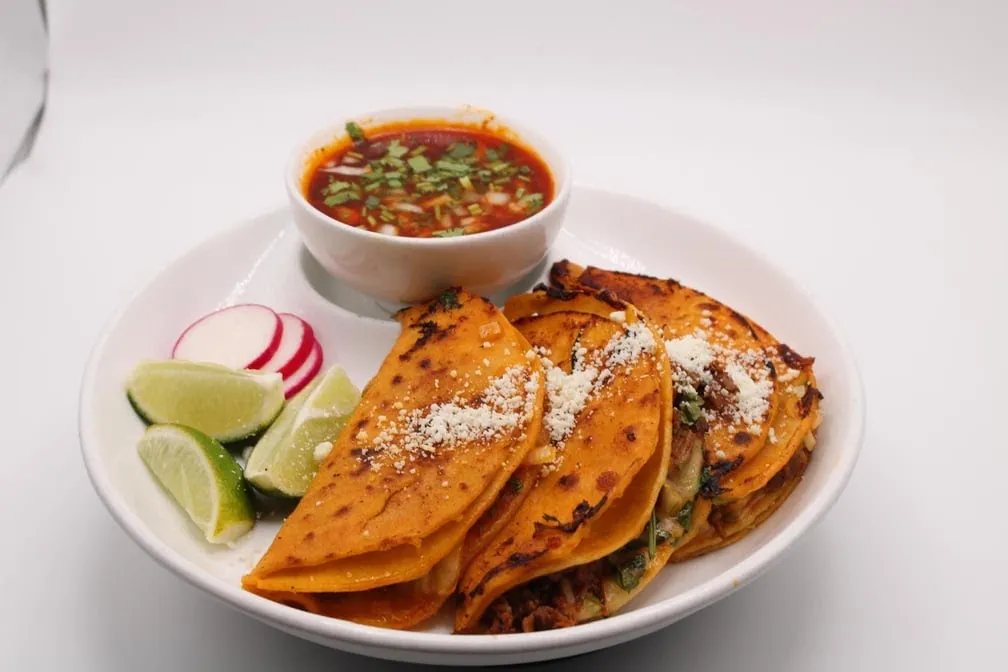 Birria tacos topped with cotija cheese, with a cup of consommé, lime and radishes
