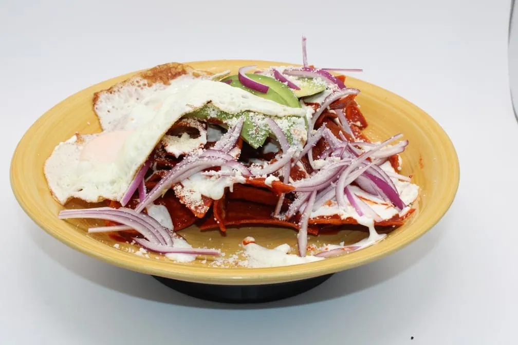 Chilaquiles rojos topped with a fried egg, avocado, red onion and crema in a yellow bowl