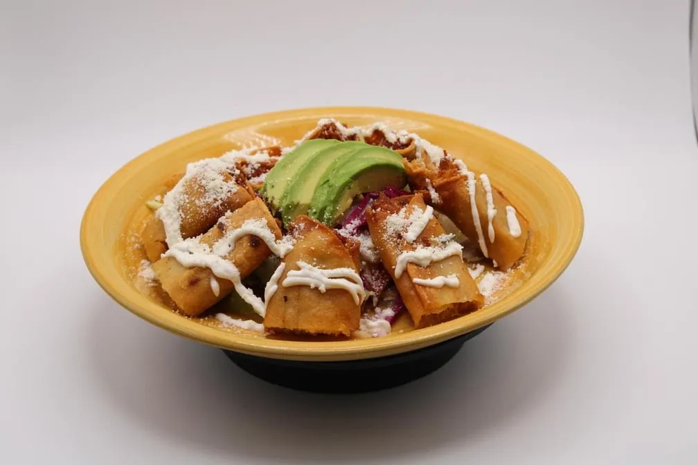 Flautas topped with crema, queso fresco and sliced avocado in a yellow bowl